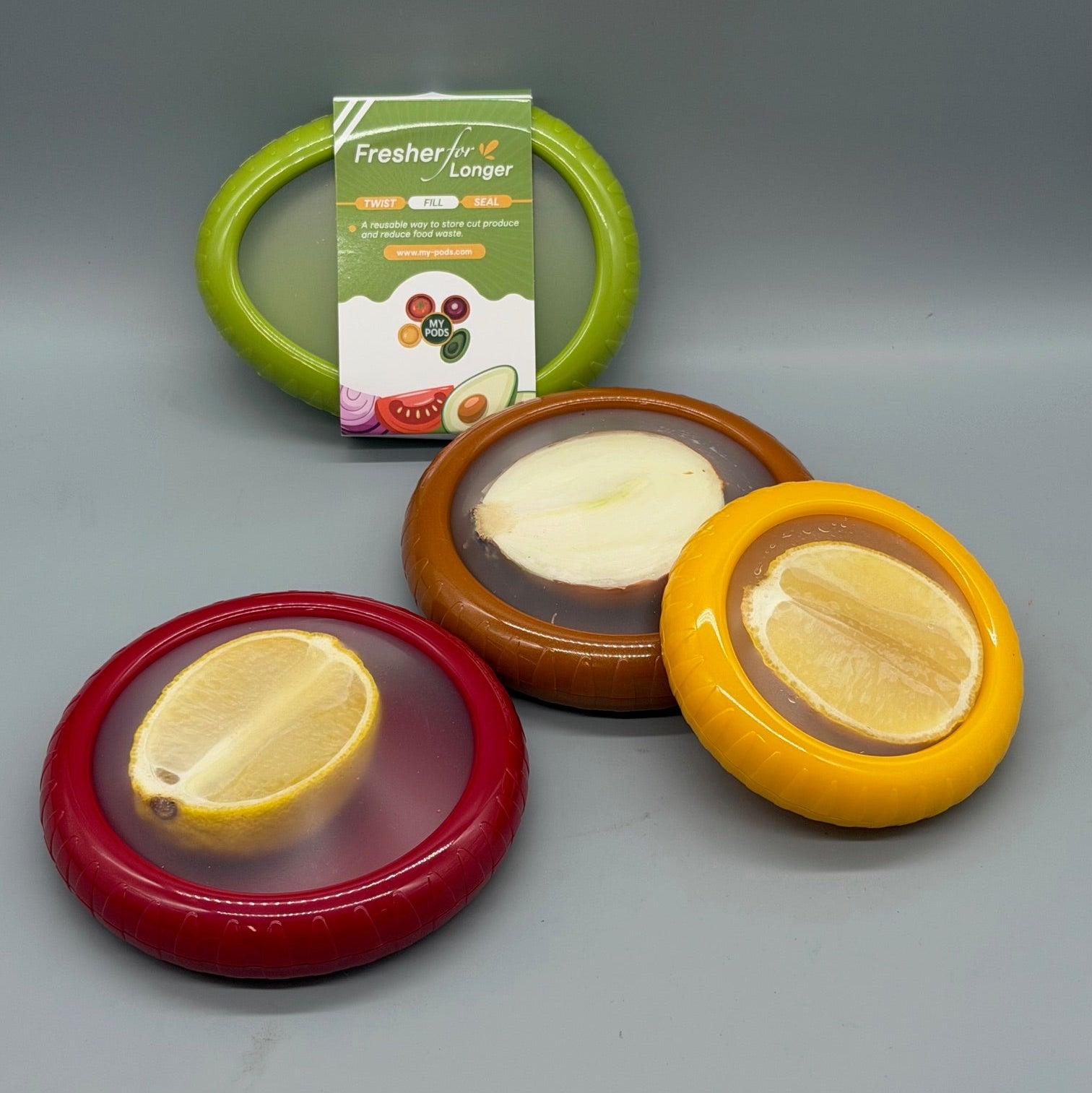 Four round fruit-preserving lids in red, brown, yellow, and green, arranged on a gray background with a product card behind the green lid. The card reads 'Freshier for Longer'.